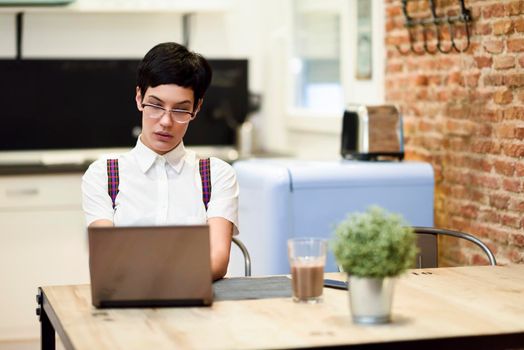 Spain, Madrid, Madrid. Young woman with very short haircut and eyeglasses typing with a laptop at home. Working at home concept.