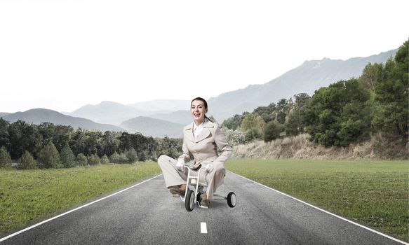 Beautiful happy woman riding children's bicycle on asphalt road. Young employee in white business suit cycling small bike outdoor. Nature landscape with green grass. Professional career start concept