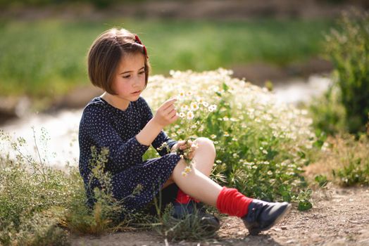 Little girl sitting in nature field wearing beautiful dress with flowers in her hand.