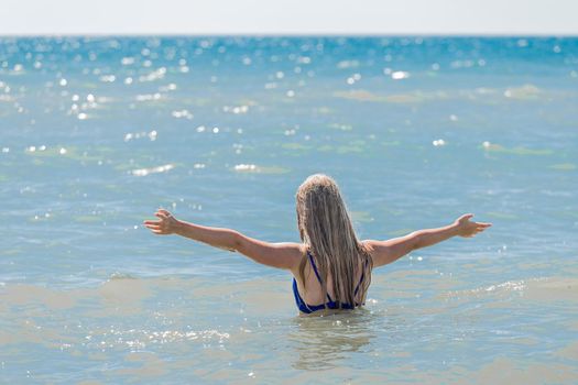 A young teenage girl with long blonde hair raised and spread her arms to the sides against the background of the sea water and horizon line. The concept of freedom.