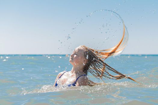 Young attractive girl teenager of European appearance throws wet, long hair up into the sea against the background of the horizon line and blue sky.