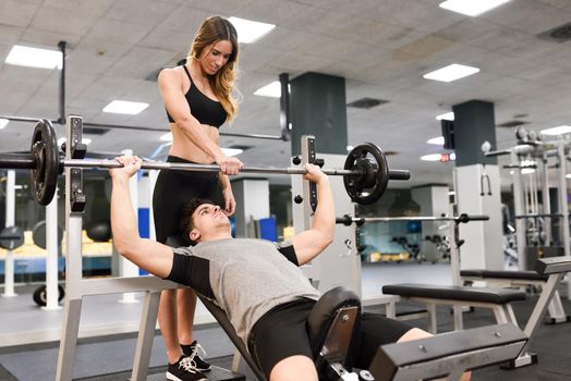 Female personal trainer helping a young man lift weights while working out in a gym