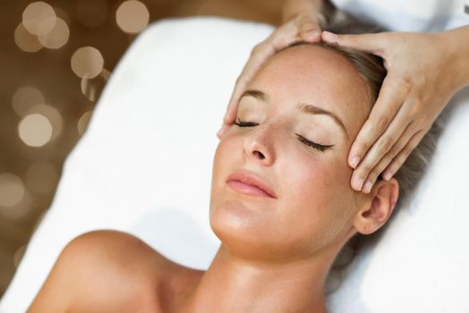 Young blond woman receiving a head massage in a spa center with eyes closed. Female patient is receiving treatment by professional therapist.