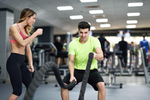 Female personal trainer motivating young man with battle ropes exercise in the fitness gym.