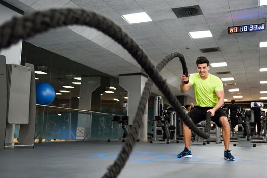 Man with battle ropes exercise in the fitness gym. Young male wearing sportswear.