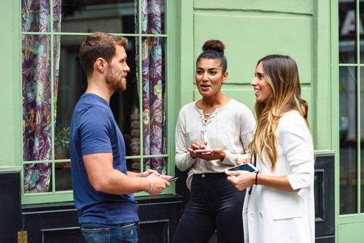 Three multiethnic people talking and smiling outdoors with smart phone in their hands. Multiracial group of friends in urban background.