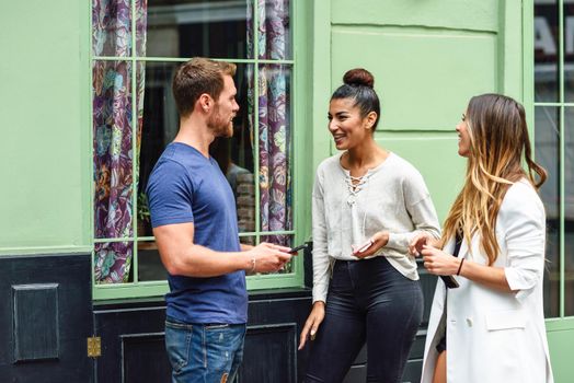 Multiracial group of friends in urban background. Three multiethnic people talking and smiling outdoors with smart phone in their hands.