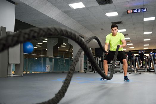 Man with battle ropes exercise in the fitness gym. Young male wearing sportswear.