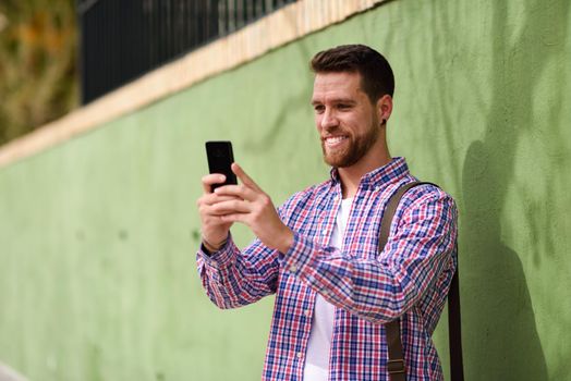 Young man looking at his smart phone and smiling in urban background. Guy wearing casual clothes. Lifestyle concept.