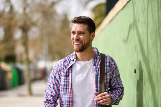 Young bearded man smiling in urban background. Traveler wearing casual clothes. Lifestyle concept.