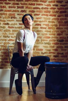 Beautiful young woman, model of fashion, with very short haircut, looking at camera sitting on chair, with a brick wall in the background. Beauty and hairstyles concept.