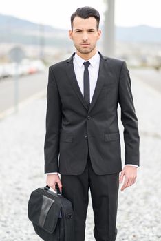 Portrait of a young businessman near a office building wearing black suit and tie with modern haircut