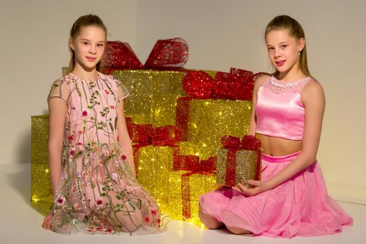 Adorable Sisters Sitting on Floor on Background Piled Gift Boxes with Red Ribbons, Portrait of Beautiful Teen Girls Celebrating Xmas and New Year Holidays, Twin Sisters Posing in Fashionable Clothes