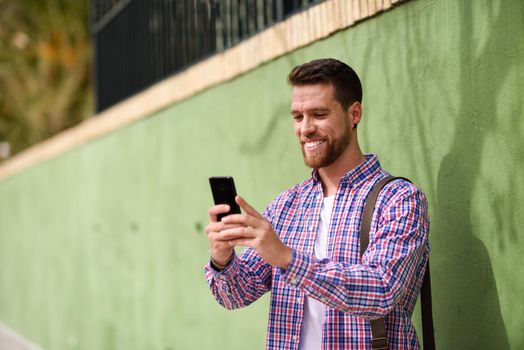 Young man looking at his smart phone and smiling in urban background. Guy wearing casual clothes. Lifestyle concept.