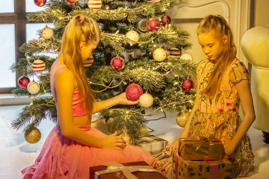 Beautiful Smiling Sisters Sitting on their Knees Opposite Each Other at Christmas Tree, Two Adorable Teen Girls Wearing Nice Dresses Exchanging Gifts at Christmas Warm Cozy Room Interior.