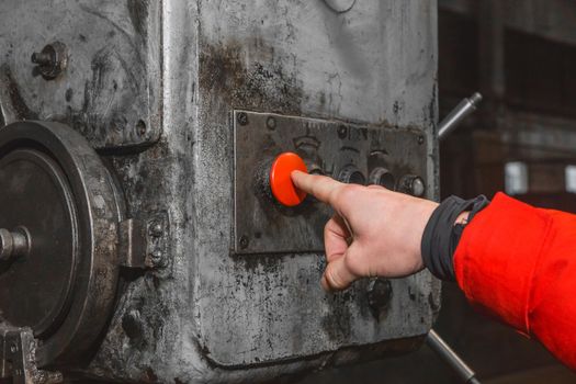 The hand of a man in a work suit presses his finger on the red button to control old industrial equipment in the factory.