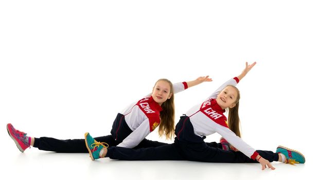 Twin Sisters Stretching Sitting on Floor, Two Smiling Girls in Sportswear and Sneakers Sitting in Horizontal Twine, Portrait of Cheerful Teenagers Doing Acrobatic Exercise Isolated on White Background