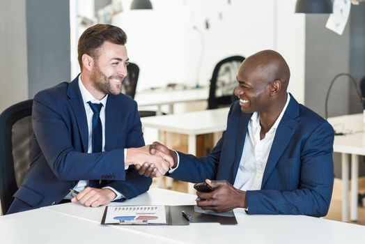 Black businessman shaking hands with a caucasian one wearing suit in a office. Two smiling men wearing blue suits working in an office with white furniture