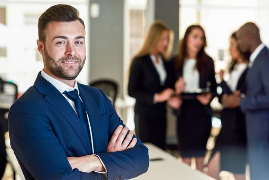 Caucasian businessman leader looking at camera in modern office with multi-ethnic businesspeople working at the background. Teamwork concept. Young man with beard wearing blue suit.