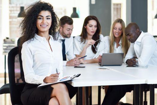 Multi-ethnic group of five businesspeople meeting in a modern office. Muslim businesswoman leader, wearing white shirt and black skirt, looking at camera.