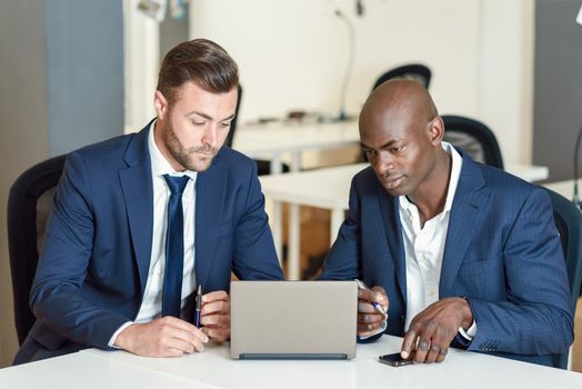 Black and caucasian businessmen looking at a laptop computer. Two men wearing blue suits working in an office with white furniture