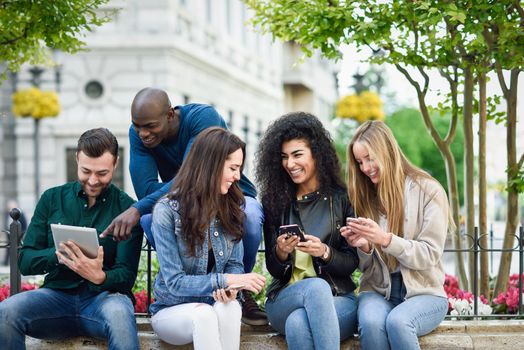 Multi-ethnic group of young people using smartphone and tablet computers outdoors in urban background. Women and men smiling and laughing in the street wearing casual clothes.