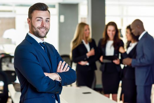 Caucasian businessman leader looking at camera in modern office with multi-ethnic businesspeople working at the background. Teamwork concept. Young man with beard wearing blue suit.