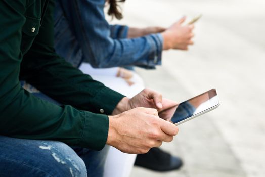 Hands of young unrecognizable people using smartphone and tablet computers outdoors in urban background. Women and men sitting on a bench in the street wearing casual clothes.