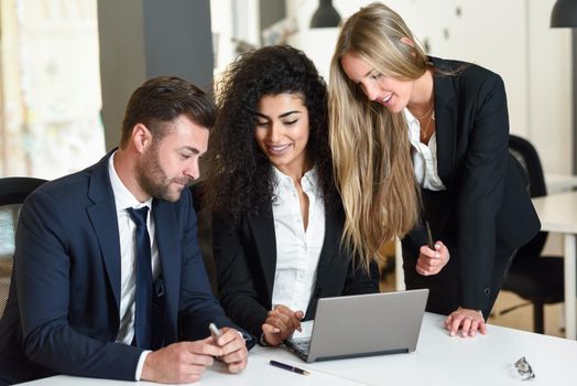 Multi-ethnic group of three businesspeople meeting in a modern office. Two women and a man wearing suit looking at a laptop computer.