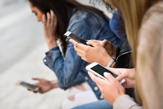 Hands of young unrecognizable people using smartphone and tablet computers outdoors in urban background. Women and men sitting on a bench in the street wearing casual clothes.