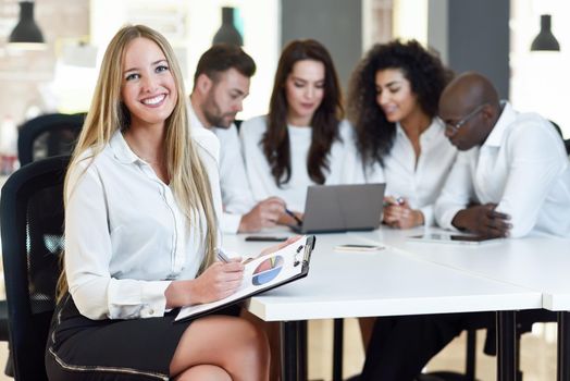 Multi-ethnic group of five businesspeople meeting in a modern office. Caucasian blonde businesswoman leader, wearing white shirt and black skirt, looking at camera.