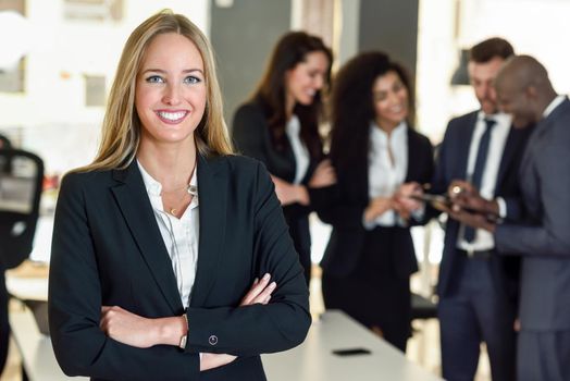 Businesswoman leader looking at camera in modern office with multi-ethnic businesspeople working at the background. Teamwork concept. Blonde caucasian woman