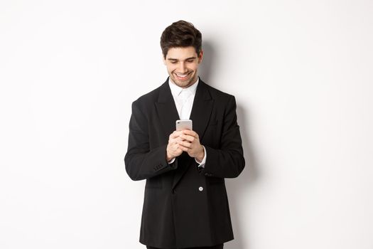 Portrait of handsome stylish businessman in black suit, writing a message, smiling and looking at smartphone, standing over white background.