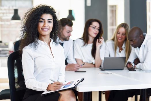 Multi-ethnic group of five businesspeople meeting in a modern office. Muslim businesswoman leader, wearing white shirt and black skirt, looking at camera.