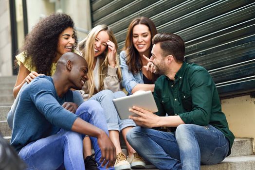 Multi-ethnic group of young people looking at a tablet computer outdoors in urban background. Group of men and woman sitting together on steps.