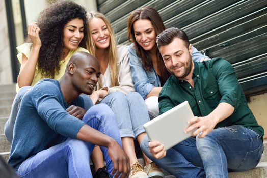 Multi-ethnic group of young people looking at a tablet computer outdoors in urban background. Group of men and woman sitting together on steps.