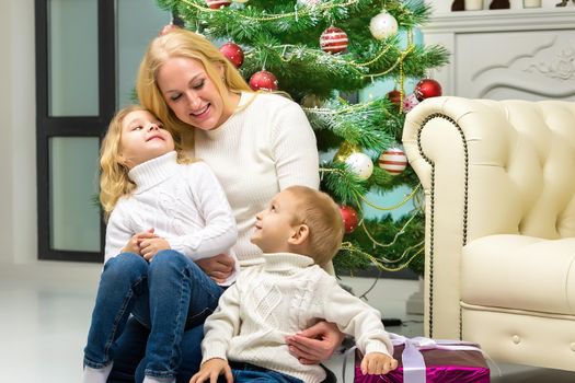 Cheerful Mom Hugging her Cute Little Kids while Sitting on the Floor in Front of Decorated Christmas Tree, Happy Family Having Fun Celebrating Xmas and New Year, Winter Holidays