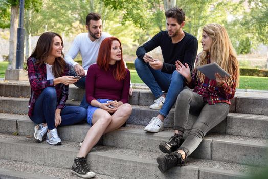 Group of young people with smartphone and tablet computers outdoors in urban background. Women and men talking on stairs in the street wearing casual clothes.