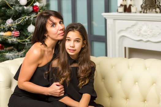 Happy mother and daughter near the Christmas tree are sitting on the couch. Family holidays concept.