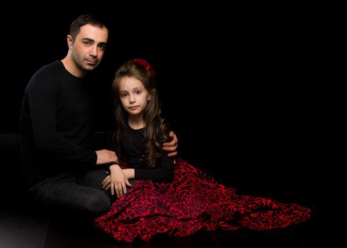 Portrait of Loving Father Hugging His Beautiful Daughter, Handsome Young Man Sitting on the Floor with his Adorable Girl, Happy Family Sitting Against Black Studio Background