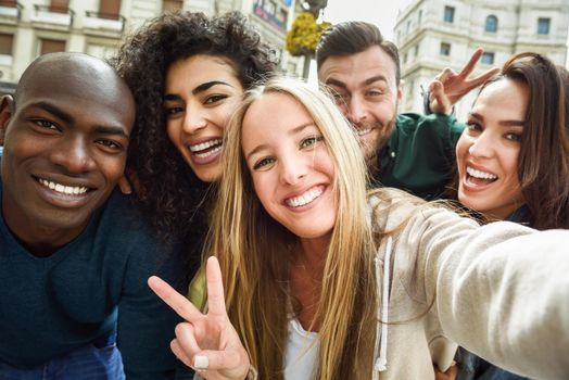 Multiracial group of friends taking selfie in a urban street with a blonde woman in foreground. Three young women and two men wearing casual clothes.