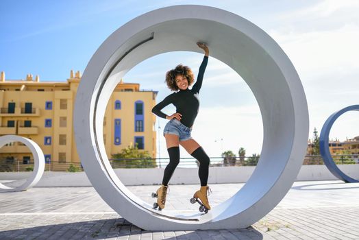 Young fit black woman on roller skates riding outdoors on urban street. Smiling girl with afro hairstyle rollerblading on sunny day