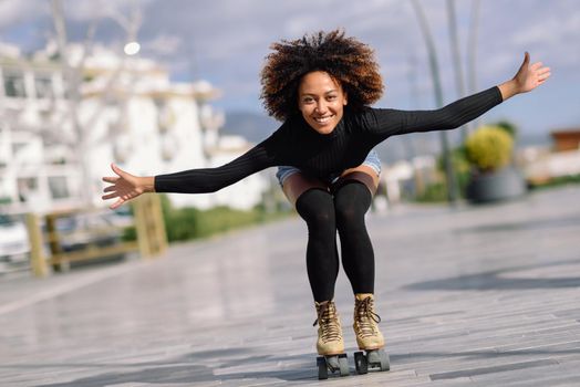 Young fit black woman on roller skates riding outdoors on urban street with open arms. Smiling girl with afro hairstyle rollerblading on sunny day