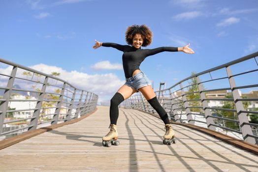 Black woman, afro hairstyle, on roller skates riding outdoors on urban bridge with open arms. Smiling young female rollerblading on sunny day. Beautiful clouds in the sky.