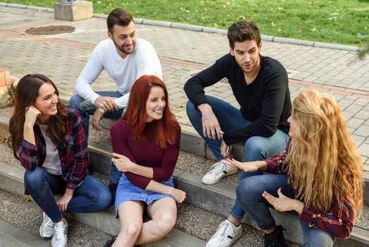 Group of young people together outdoors in urban background. Women and men sitting on stairs in the street wearing casual clothes.