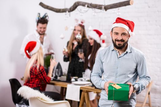 Shot of happy friends enjoying holidays. Focus on the man in the foreground in a red Christmas hat. A man with a gift in his hands in a white room