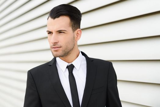 Portrait of a young businessman near a office building wearing black suit and tie with modern haircut