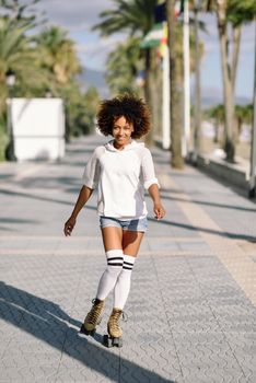 Smiling black woman on roller skates riding outdoors on beach promenade with palm trees. Smiling girl with afro hairstyle rollerblading on sunny day.