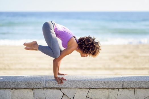 Black woman, afro hairstyle, doing yoga in the beach. Young Female wearing sport clothes with sea at the background