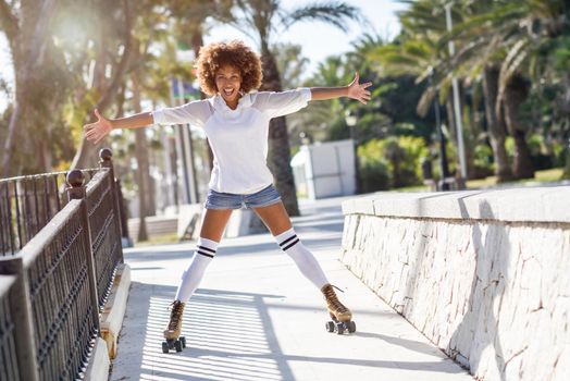 Young black woman on roller skates riding near the beach. Girl with afro hairstyle rollerblading on sunny promenade with sun backlight.